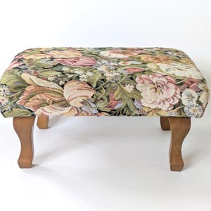 Könnte beinhalten: Ein rechteckiger Fußhocker mit einem gewebten Gobelin-Bezug, der ein dichtes Blumenmuster in Rosa-, Creme-, Grün- und Schwarztönen aufweist. Das Möbelstück steht auf vier hellbraunen Holzbeinen mit geschwungenem Cabriole-Design.