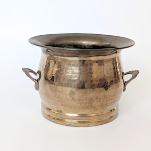 Puede incluir: Una jardinera de latón vintage con textura martillada y dos asas ornamentadas. La jardinera tiene un borde ancho y oscuro y un cuerpo redondeado. La base tiene una banda decorativa. Adecuada para uso en interiores o exteriores. Mide unos 20 cm de diámetro.