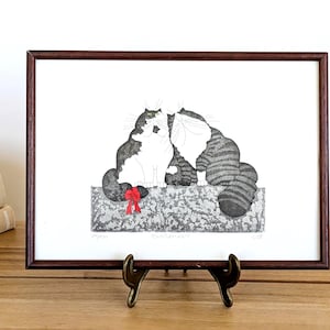 May include: A framed art print featuring two stylised cats with black and white striped fur sitting together on a textured grey surface. One cat has a bright red bow tied to its tail. The print is enclosed in a dark brown wooden frame.