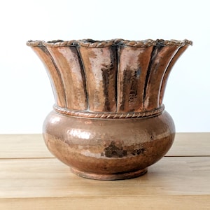 Puede incluir: Un jarrón de cobre martillado con un borde acanalado y una base bulbosa. El jarrón tiene un detalle en forma de cuerda alrededor del centro y un color marrón cálido. El cobre tiene una superficie reflectante y el jarrón está sobre una superficie de madera.