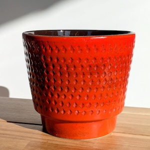Könnte beinhalten: Ein leuchtend roter Keramik-Blumentopf mit einem strukturierten Rautenmuster. Der Topf hat eine breite, leicht ausgestellte Form und einen dunkelbraunen Innenraum. Er steht auf einem kleinen, erhöhten Sockel, ideal für Pflanzen. Der Topf steht auf einer Holzoberfläche.