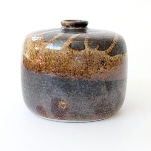 Könnte beinhalten: Eine Keramikvase mit rundem Körper und kleiner Öffnung. Die Vase hat eine Farbpalette aus Braun, Beige und Dunkelgrau. Der obere Teil hat ein strukturiertes, gesprenkeltes Aussehen, während der untere Abschnitt einen dunkleren, gesprenkelten Farbton aufweist.