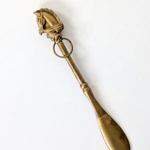 Könnte beinhalten: Ein Schuhlöffel aus Messing mit einem geformten Pferdekopfgriff und einem kleinen befestigten Metallring. Der lange, schlanke Schaft führt zu einem glatten, gebogenen Löffelende. Das Metall hat einen warmen, gealterten Goldton mit Patina.
