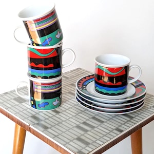 Peut inclure: Ensemble de tasses à café et soucoupes vintage avec un motif géométrique coloré. Les tasses sont empilées sur une petite table à surface carrelée. Les tasses et soucoupes présentent un fond noir avec des accents rouges, bleus et verts.
