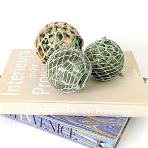 Puede incluir: Tres bolas decorativas de vidrio verde, envueltas en redes, descansan sobre dos libros. Una bola está en una red beige, las otras en redes blancas. Los libros tienen cubiertas beige con texto en alemán e inglés.