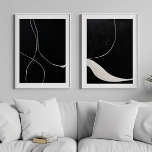 K&ouml;nnte beinhalten: Zwei gerahmte abstrakte Kunstwerke. Jedes hat einen schwarzen Hintergrund mit wei&szlig;en, flie&szlig;enden Linien. Die wei&szlig;en Rahmen kontrastieren mit der Kunst, die &uuml;ber einem wei&szlig;en Sofa mit Kissen ausgestellt ist. Der Stil ist modern und minimalistisch.