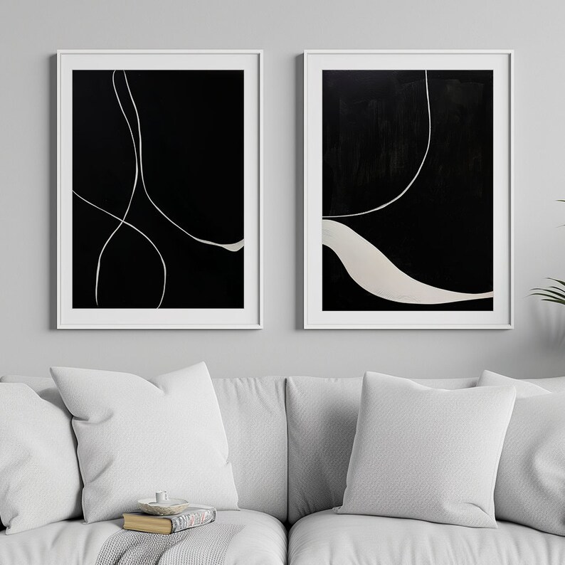 K&ouml;nnte beinhalten: Zwei gerahmte abstrakte Kunstwerke. Jedes hat einen schwarzen Hintergrund mit wei&szlig;en, flie&szlig;enden Linien. Die wei&szlig;en Rahmen kontrastieren mit der Kunst, die &uuml;ber einem wei&szlig;en Sofa mit Kissen ausgestellt ist. Der Stil ist modern und minimalistisch.