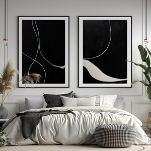 K&ouml;nnte beinhalten: Zwei gerahmte abstrakte Kunstwerke. Jedes hat einen schwarzen Hintergrund mit wei&szlig;en, geschwungenen Linien. Die Kunstwerke werden &uuml;ber einem Bett mit einer wei&szlig;en Bettdecke, grauen Kissen und einem gemusterten Hocker ausgestellt. Der Raum ist mit Pflanzen und neutralen T&ouml;nen dekoriert.