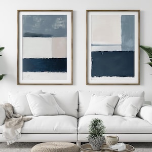 Op de afbeelding: Twee abstracte schilderijen in houten frames met een blauw, wit en roze kleurenschema. De schilderijen hangen aan een witte muur boven een witte bank.