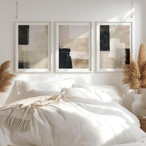 Op de afbeelding: Drie abstracte kunstprints in witte frames met een beige en zwart kleurenschema. De prints hangen boven een wit bed met witte beddengoed en een beige deken.
