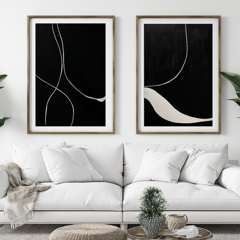 K&ouml;nnte beinhalten: Zwei gerahmte abstrakte Kunstdrucke. Jeder hat einen schwarzen Hintergrund mit wei&szlig;en, geschwungenen Linien. Die Rahmen sind warmbraun. Das Kunstwerk wird &uuml;ber einem wei&szlig;en Sofa mit Kissen pr&auml;sentiert und schafft eine moderne &Auml;sthetik.