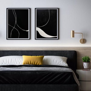 K&ouml;nnte beinhalten: Zwei gerahmte Kunstdrucke mit schwarzen Hintergr&uuml;nden und wei&szlig;en Linienmustern h&auml;ngen &uuml;ber einem Bett. Das Bett hat ein schwarzes Kopfteil, eine schwarze Bettdecke und wei&szlig;e und gelbe Kissen. Eine goldene Wandleuchte und eine kleine Topfpflanze sind ebenfalls zu sehen.