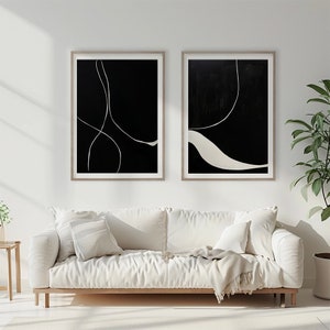 K&ouml;nnte beinhalten: Zwei gerahmte abstrakte Kunstdrucke. Jeder Druck hat einen schwarzen Hintergrund mit wei&szlig;en Linien und Formen. Die Rahmen sind hellholzfarben. Das Kunstwerk wird &uuml;ber einem wei&szlig;en Sofa mit Kissen und einer Decke ausgestellt.
