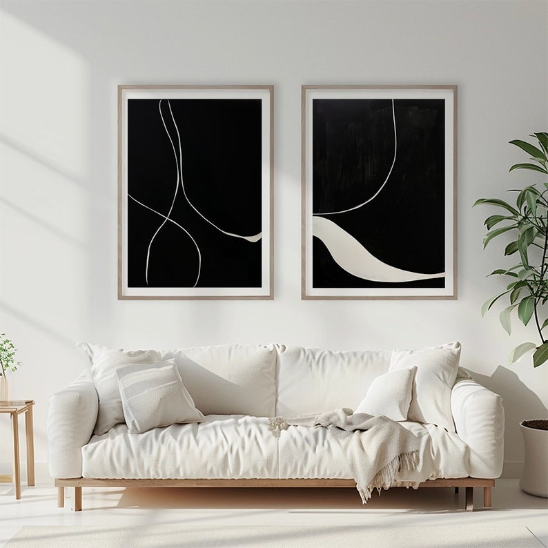 K&ouml;nnte beinhalten: Zwei gerahmte abstrakte Kunstdrucke. Jeder Druck hat einen schwarzen Hintergrund mit wei&szlig;en Linien und Formen. Die Rahmen sind hellholzfarben. Das Kunstwerk wird &uuml;ber einem wei&szlig;en Sofa mit Kissen und einer Decke ausgestellt.