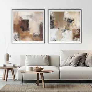 Può includere: Due dipinti astratti in tonalità marrone, beige e bianco con accenti neri. I dipinti sono incorniciati in nero e appesi a una parete bianca sopra un divano bianco.