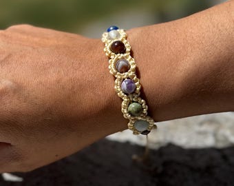 May include: A beige macrame bracelet with a variety of colorful gemstone beads. The beads are round and have a natural, polished finish. The bracelet is tied with a knot at the end.