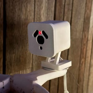 May include: A white, cube-shaped security camera with a red light and a black lens. The camera is mounted on a white bracket and is attached to a white arm. The background is a wooden wall.