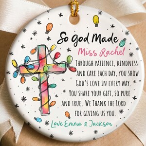 May include: A round, white ceramic Christmas ornament with a gold string for hanging. The ornament features a colorful cross decorated with lights and text that reads "So god Made Miss Rachel". The ornament is decorated with snowflakes and light bulbs.