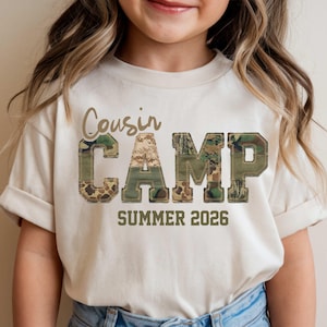 May include: A cream-colored t-shirt with the words "Cousin Camp" in a brown script font. The word "CAMP" is in large, block letters with a camouflage pattern. Below, the text "SUMMER 2026" is printed in a brown font.