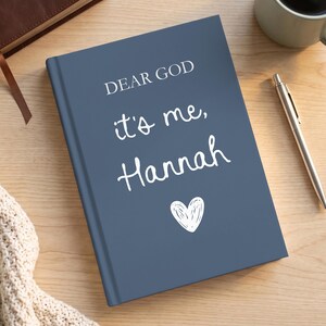 Puede incluir: Un diario de tapa dura color azul pizarra con el texto impreso en blanco "DEAR GOD it's me, Hannah" y un pequeño icono de corazón blanco dibujado a mano centrado debajo del texto. El diario está sobre una superficie de madera clara junto a un bolígrafo plateado.
