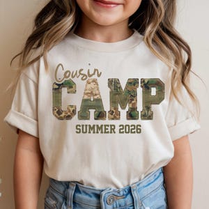 May include: A cream-colored short-sleeve t-shirt featuring the text Cousin CAMP in large block letters with a camouflage pattern and a stitched border effect. Below the word CAMP, the text SUMMER 2026 is printed in a dark olive green font.