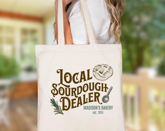 May include: A cream-colored canvas tote bag featuring the brown text Local Sourdough Dealer, a graphic of a loaf of bread, a small spoon, and a sprig of rosemary. Below the text, it reads Madison's Bakery est. 2024 in a smaller green font.