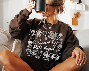 May include: A dark chocolate brown long-sleeve sweatshirt featuring white graphic illustrations of speech therapy icons, including alphabet blocks, a magnifying glass, a brain, and a stethoscope, with the text Speech Pathologist in the center.