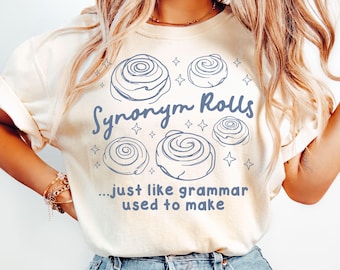 May include: An ivory t-shirt featuring a blue graphic of four cinnamon rolls surrounded by small star icons. The text reads "Synonym Rolls" above the rolls and "...just like grammar used to make" below the graphic in a blue sans-serif font.