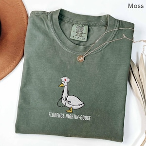 May include: A moss green t-shirt featuring an embroidered white goose wearing a nurse hat with a red cross and a stethoscope. Below the goose, the text reads FLORENCE NIGHTIN-GOOSE. The shirt has a Comfort Colors label inside the collar.