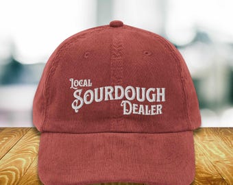 May include: A brick red corduroy baseball cap featuring the white embroidered text Local SOURDOUGH DEALER on the front panel. The hat has a structured crown, a curved brim, and a button on top, showcasing a classic six-panel design with stitching.