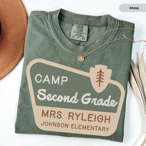 May include: A moss green t-shirt featuring the text "CAMP Second Grade MRS. RYLEIGH JOHNSON ELEMENTARY". The shirt has a relaxed fit and is made of a soft material. A gold necklace is draped across the shirt.