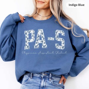 May include: An indigo blue crewneck sweatshirt featuring the text PA-S in a collegiate font with a white and blue floral pattern. Below the large letters, the text Physician Assistant Student is printed in a thin, white cursive script.
