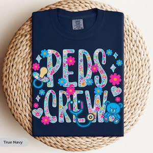 May include: A navy blue t-shirt featuring the text PEDS CREW in large letters filled with a colorful floral pattern. The design includes a pacifier, a stethoscope, a teddy bear, hearts, and small flowers in shades of pink, blue, and white.