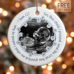 May include: A round white ceramic ornament featuring a central black and white ultrasound image of a fetus. The image is encircled by a blue floral vine pattern and the text, For this child I prayed, and the Lord has granted me what I asked of him. - 1 Sam 1:27.