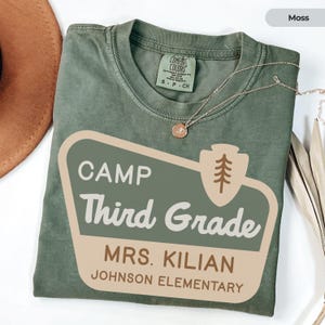 May include: A moss green t-shirt with the text "CAMP Third Grade MRS. KILIAN JOHNSON ELEMENTARY". The shirt has a crew neck and short sleeves. A gold necklace with a pendant is draped across the shirt.