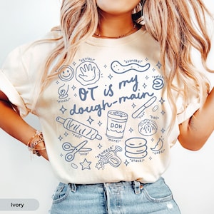 May include: An ivory t-shirt featuring blue line art illustrations of play dough tools and actions. The text reads OT is my dough-main, with labels including squish, sssnake, pinch, twist, cut, ball, roll, more, snip, squeeze, and stack.