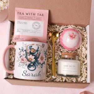 May include: Gift box with a white mug featuring a stethoscope and floral design, personalized with the name "Sarah." Includes tea, a bath bomb, a candle, and a gold spoon. The tea packet reads "Tea With Tae."