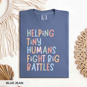 May include: A blue jean colored t-shirt with a crew neckline and a graphic print in pastel pink, yellow, and light blue. The text reads HELPING TINY HUMANS FIGHT BIG BATTLES in a playful, multi-colored sans-serif font. The shirt has a Comfort Colors label.