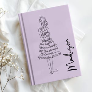 May include: A lavender journal with a black line drawing of a woman in a dress. The text on the dress reads "She is clothed with strength and dignity, she can laugh at the days to come... Proverbs 31:25." The name "Madison" is written in cursive.