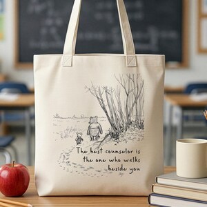 May include: Cream-colored tote bag featuring a black and white illustration of Winnie the Pooh and Piglet walking. The text "The best counselor is the one who walks beside you" is printed below the image. The bag has two cream-colored handles.