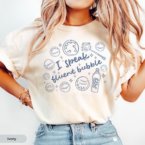 May include: An ivory t-shirt featuring a blue graphic print with the text "I speak fluent bubble" surrounded by illustrations of bubbles, a bubble wand, and a bubble solution bottle with phrases like "more!", "again!", "you did it!", "Pop!", "Your Turn!", and "Yay!".