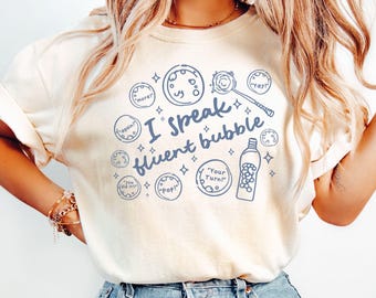 May include: An ivory t-shirt featuring a blue graphic print with the text "I speak fluent bubble" surrounded by illustrations of bubbles, a bubble wand, and a bubble solution bottle with phrases like "more!", "again!", "you did it!", "Pop!", "Your Turn!", and "Yay!".