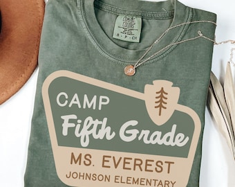 Peut inclure: Un t-shirt vert mousse avec un motif « CAMP Fifth Grade MS. EVEREST JOHNSON ELEMENTARY ». Le t-shirt a un col rond et des manches courtes. Un collier doré avec un pendentif est drapé sur le t-shirt.