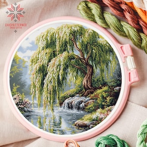May include: A cross-stitch embroidery of a weeping willow tree over a river, set in a pink embroidery hoop. The scene includes a waterfall, lush greenery, and a cloudy sky. The text "CHICPATTERNCO CROSS STITCH" is visible.