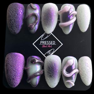 May include: A collection of ten press-on nails with diverse designs. Some nails feature textured purple, others have a pearlescent finish with a snake motif, and some are white with a droplet effect. The text "PRESSED Nail Art" is displayed.