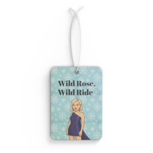 May include: Rectangular air freshener with a light blue background and a snowflake pattern. The air freshener features the text "Wild Rose, Wild Ride" and an illustration of a woman in a purple dress. A white string is attached for hanging.
