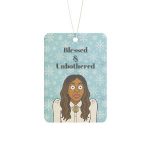 May include: Air freshener with a light blue background and snowflake pattern. The text "Blessed & Unbothered" is above a cartoon illustration of a woman with wide eyes. A white string is attached for hanging.