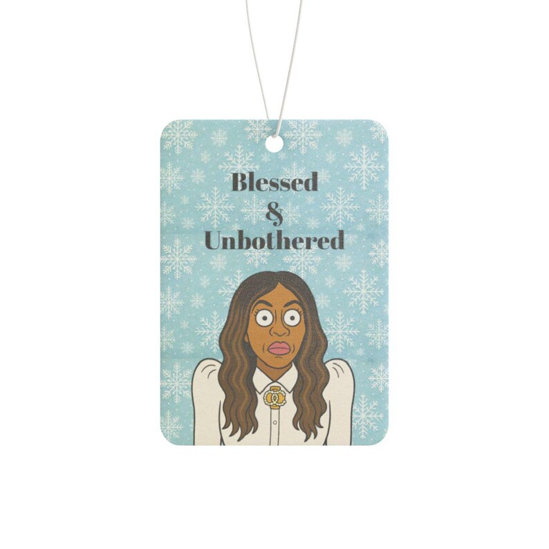 May include: Air freshener with a light blue background and snowflake pattern. The text "Blessed & Unbothered" is above a cartoon illustration of a woman with wide eyes. A white string is attached for hanging.