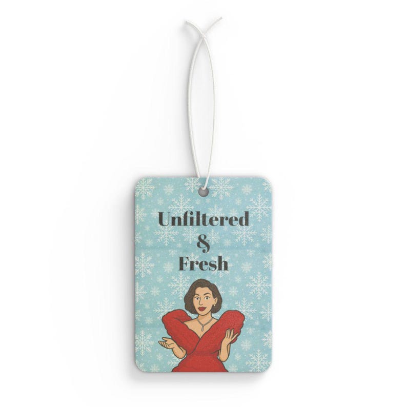 May include: Rectangular air freshener with a light blue background and white snowflakes. The air freshener has the words "Unfiltered & Fresh" and a cartoon illustration of a woman in a red dress. A white string is attached for hanging.