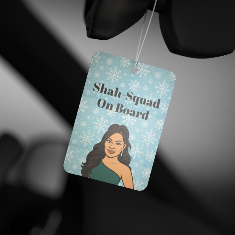 May include: A light blue car air freshener with a snowflake pattern and the text "Shah-Squad On Board." The air freshener features an illustration of a woman wearing a green dress.